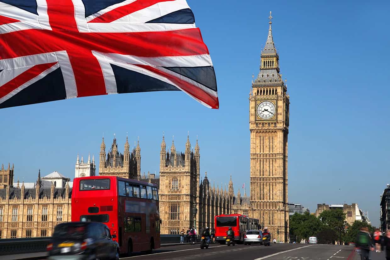 A Union Jack flapping in the wind, with Big Ben in the background and a Double Decker red London bus driving past.