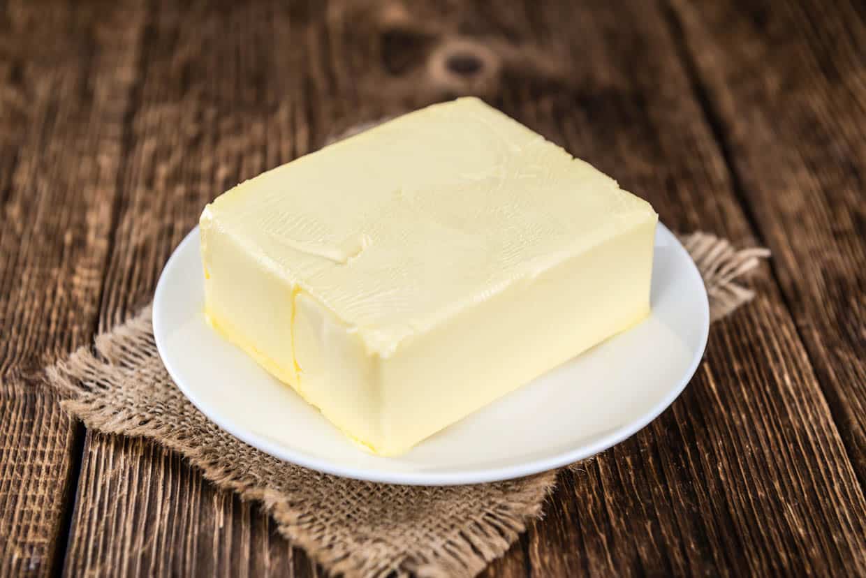 Low angle shot of a block of softened butter on a white plate on top of a square of burlap on a wooden table.