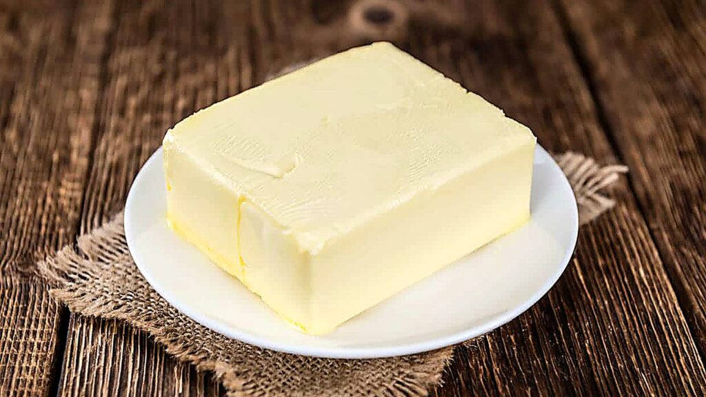 A rectangular block of butter sits on a white plate, placed on a piece of burlap over a wooden table&mdash;a perfect setup for anyone curious about how to soften butter before using it in recipes.