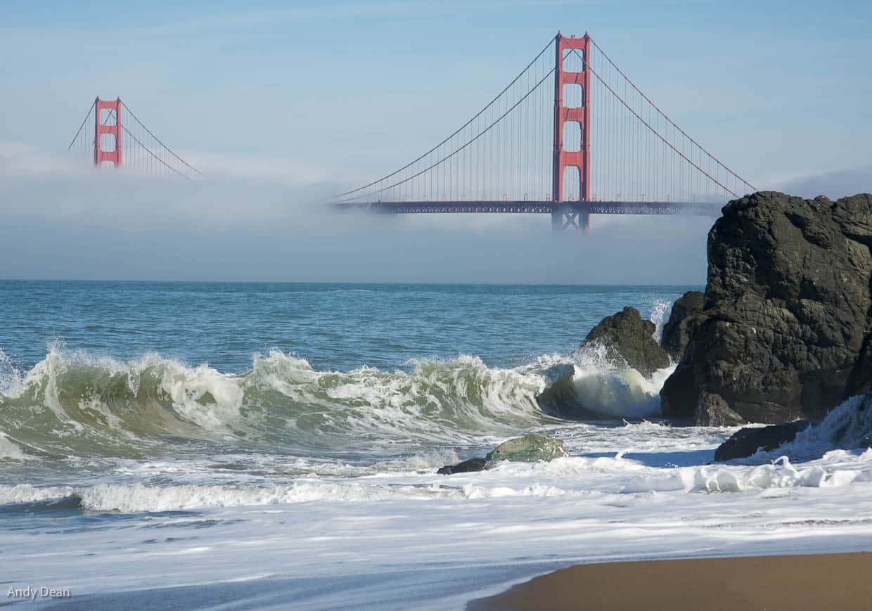 A photo of the Golden Gate Bridge peaking out of the fog, shot from Baker Beach to the West. YOu can see the sand and waves breaking on Baker Beach in the foreground.