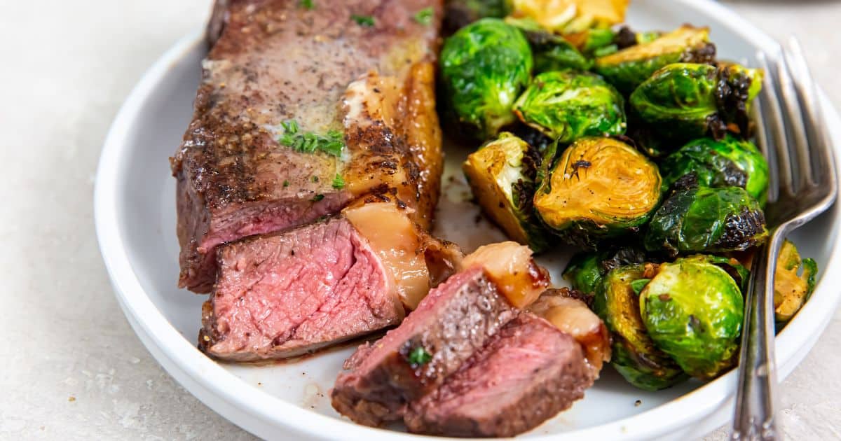 Air Fryer New York Strip Steaks on a white plate with butter, thyme, pepper, salt, brussels sprouts, and a fork.
