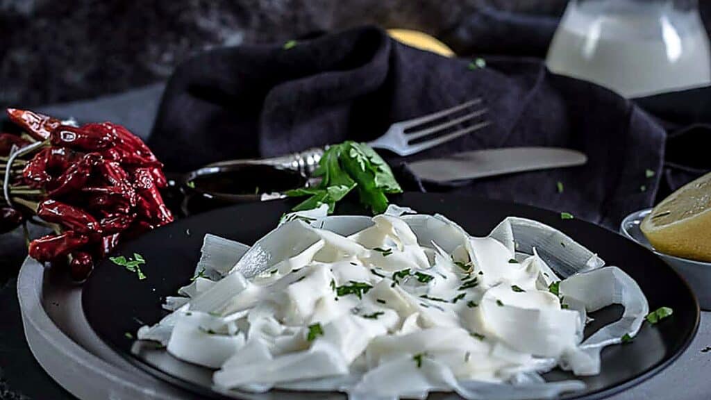 A black plate with wide, white veggie noodles ribbons garnished with herbs, placed near red chili peppers, a lemon, a fork, and a bottle of milk on a dark cloth background.