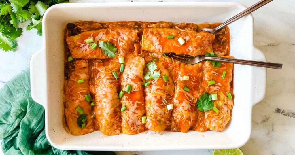 A baking dish full of chicken enchiladas is surrounded by cut limes, a halved avocado, and a green linen.
