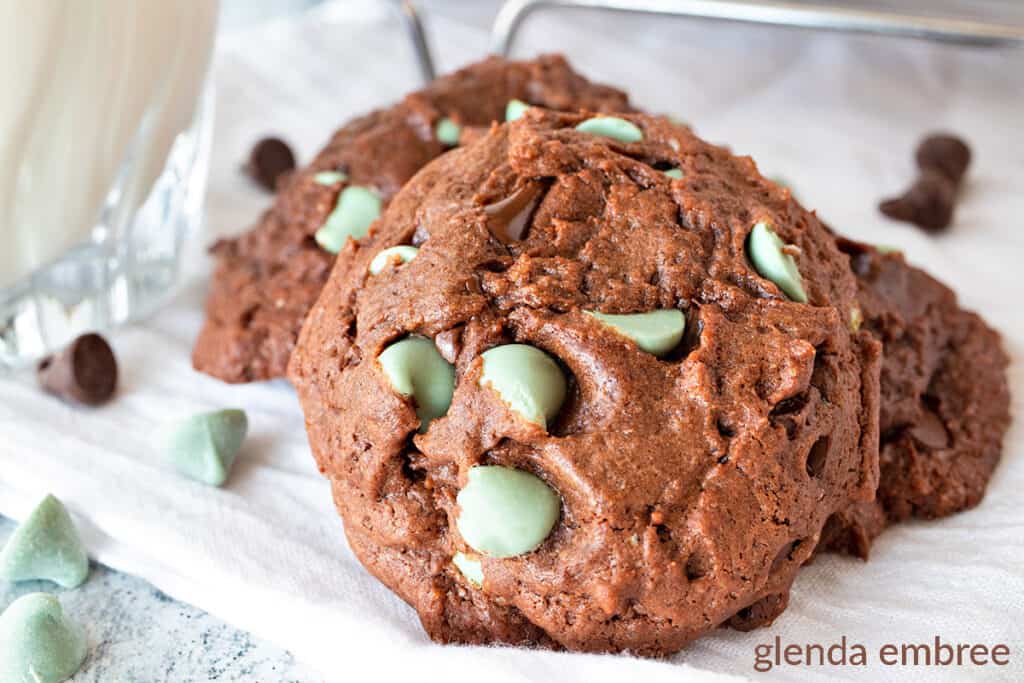 Three Chocolate Mint Chip Cookies stacked on a white muslin cloth.  Chocolate and mint chips are scattered around the cookies. 