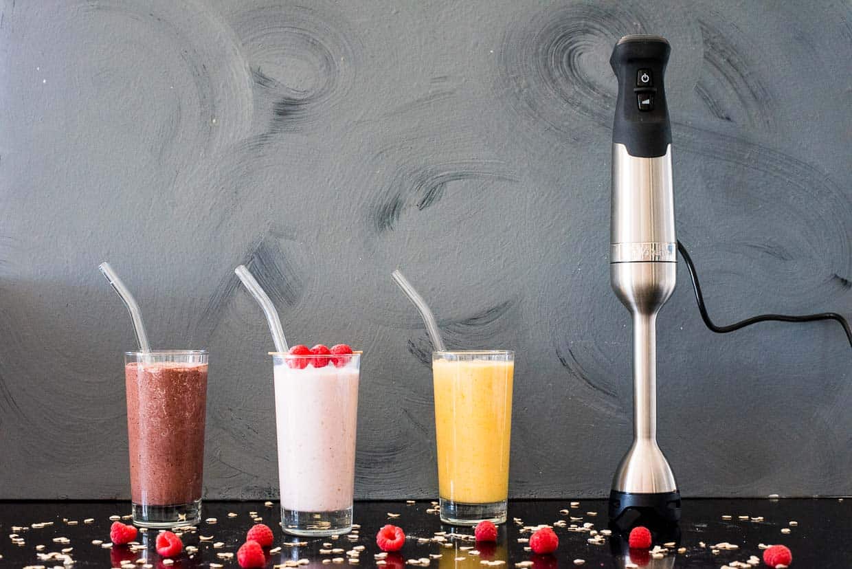 Three smoothies: chocolate cherry, raspberry banana, and peach banana, next to a Vitamix immersion blender with a gray background.