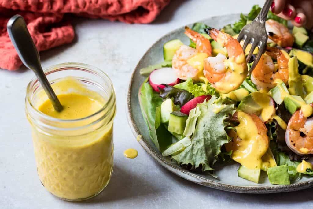 A glass jar filled with a yellow sauce sitting next to a plate of salad.