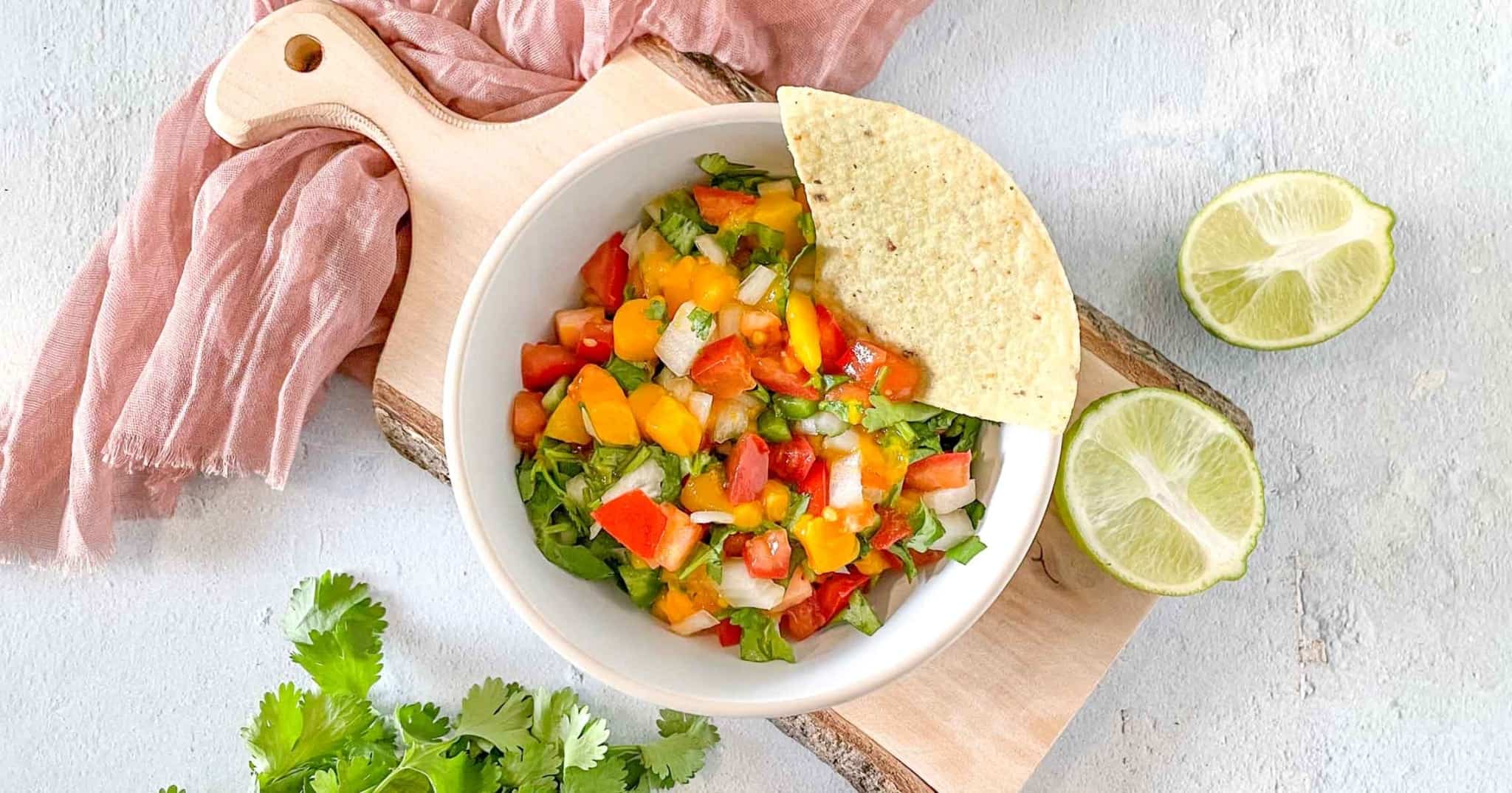 A bowl of mango pico de gallo sits on a rustic wooden cutting board surrounded by limes, cilantro, and a pink linen.