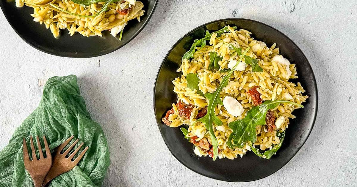 Two plates of orzo pesto salad sit on a concrete background with a green linen and copper forks.