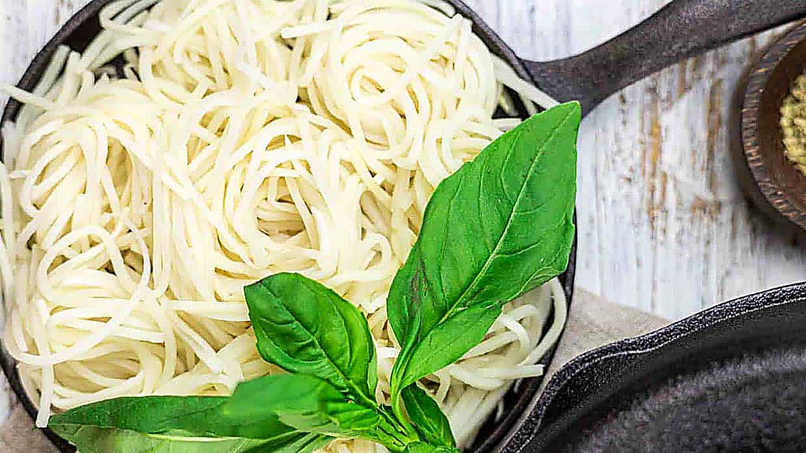 Cooked veggie noodles in a black skillet topped with fresh basil leaves, placed on a light wooden surface.