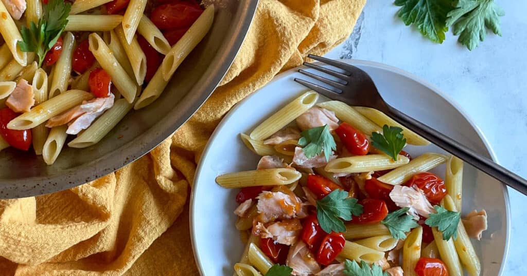 A closeup of penne pasta with burst cherry tomatoes, flaked salmon, and parsley on a white plate.