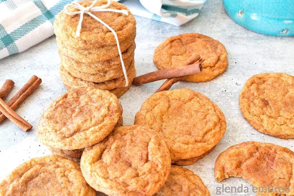 Chai Spiced Pumpkin Snickerdoodles on a concrete countertop.  A stack of five cookies is tied with a cooking twine bow.  Additional cookies and cinnamon sticks are scattered around the stack.  One cookie in the foreground has a bite out of it. 