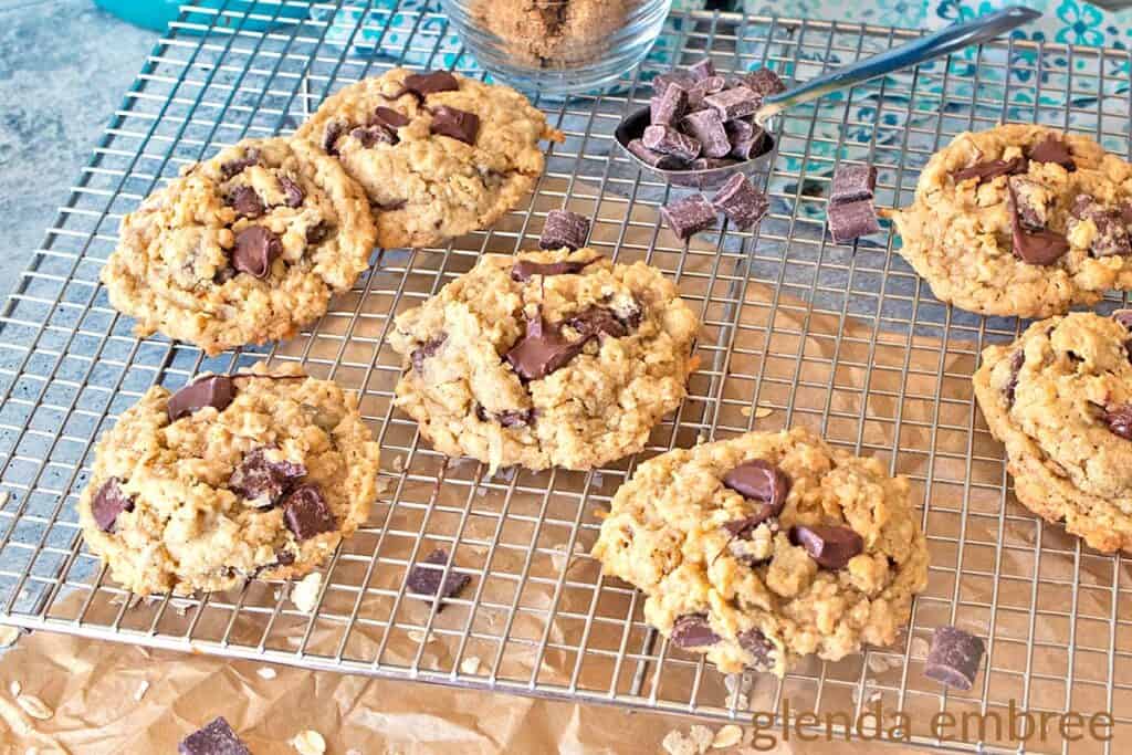 Ranger Cookies cooling on a wire baking rack.  Rack is sitting on a concrete countertop that is partially covered in brown paper.  A Tablespoon full of chocolate pieces is sitting next to the cookies on the wire rack.