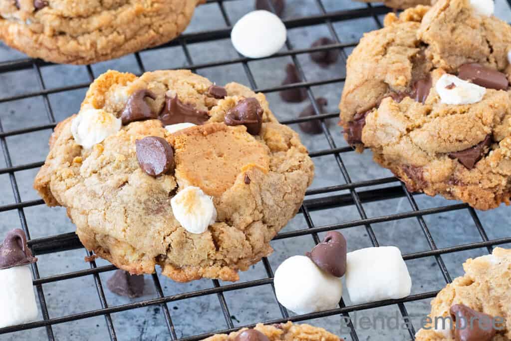 S'mores Cookies cooling on a black wire rack.  Rack is sitting on a concrete countertop and their are chocolate chips and marshmallows strewn about.