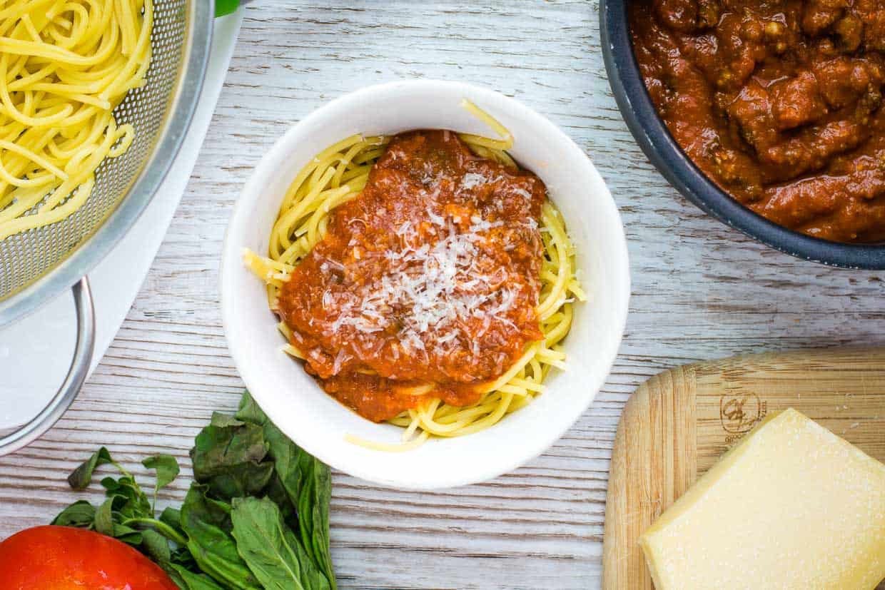 Meaty tomato sauce on a bed of spaghetti noodles in a white background, with a pan of sauce and a bowl of noodles.