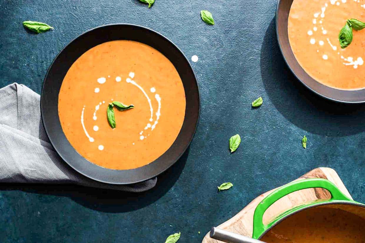 Two black bowls of tomato basil bisque on a blue background. 