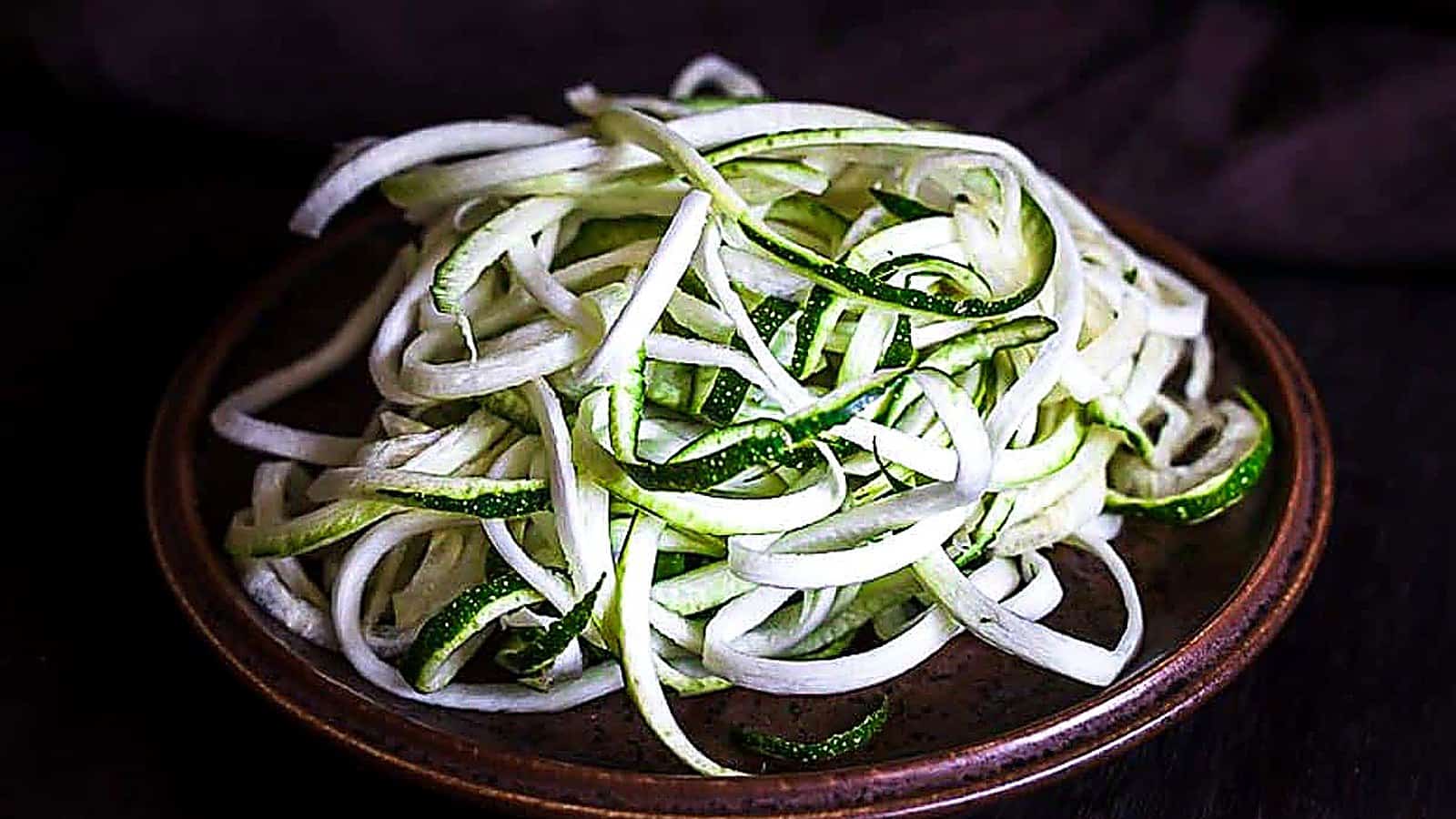A plate of raw spiralized zucchini noodles, also known as zoodles or veggie noodles, sits on a brown ceramic dish against a dark background.