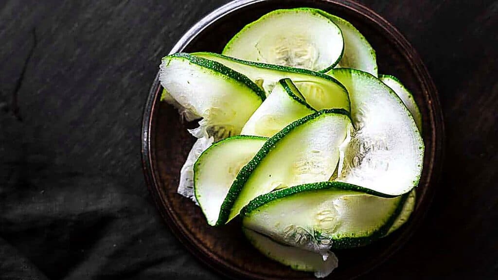 A small dark plate holds thin, overlapping slices of raw zucchini&mdash;resembling delicate veggie noodles&mdash;on a dark surface.