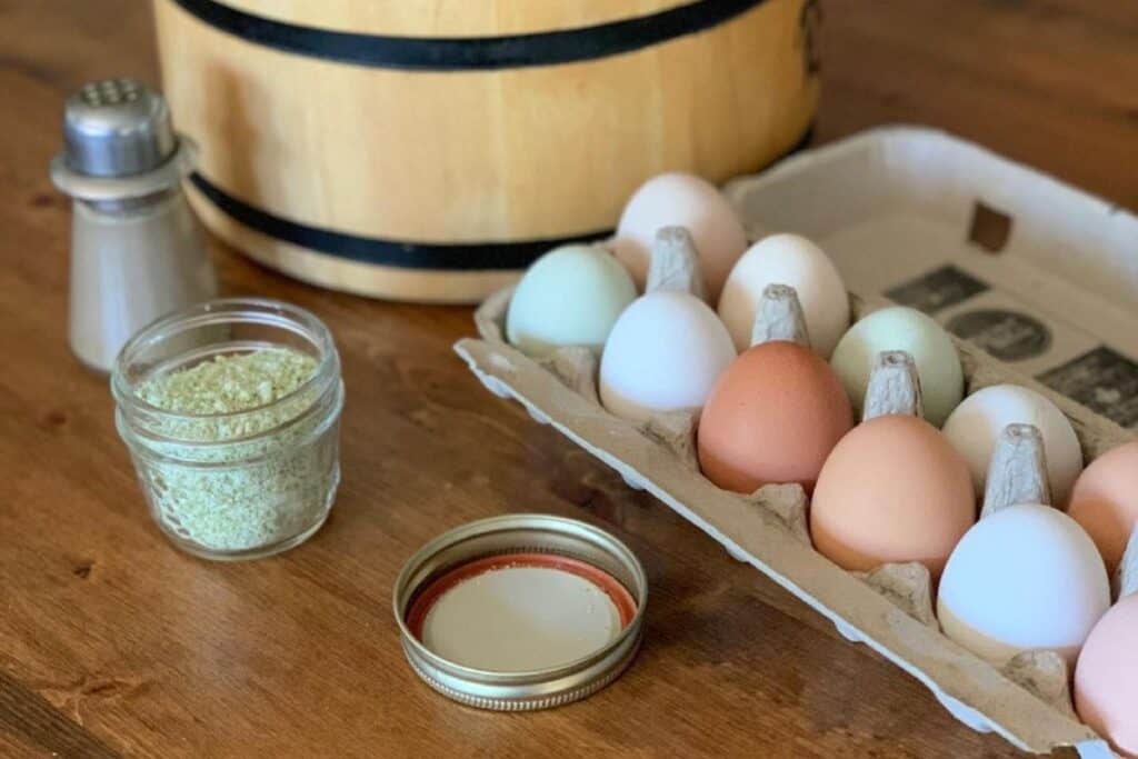 Homemade spruce tip salt in a mason jar, farm fresh eggs in carton, and wooden banded bowl on wooden counter.