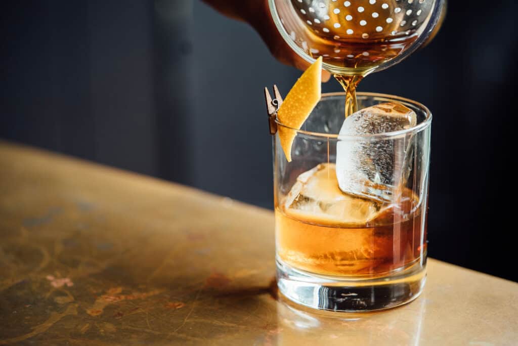 A cocktail shaker pouring whiskey through a Boston strainer into an old-fashioned cocktail.
