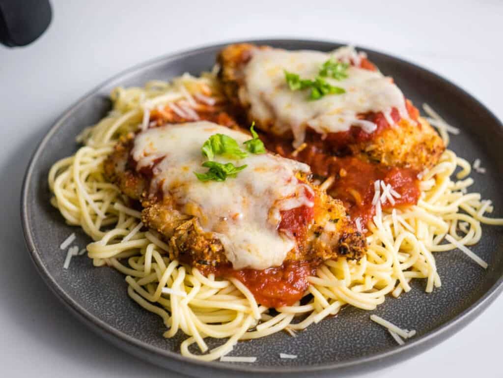 Breaded air fryer chicken breasts topped with marinara sauce and melted cheese on a bed of pasta.