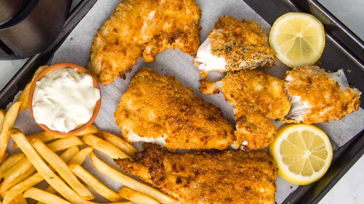 Fish and fries on a metal tray with tartar sauce and lemon.