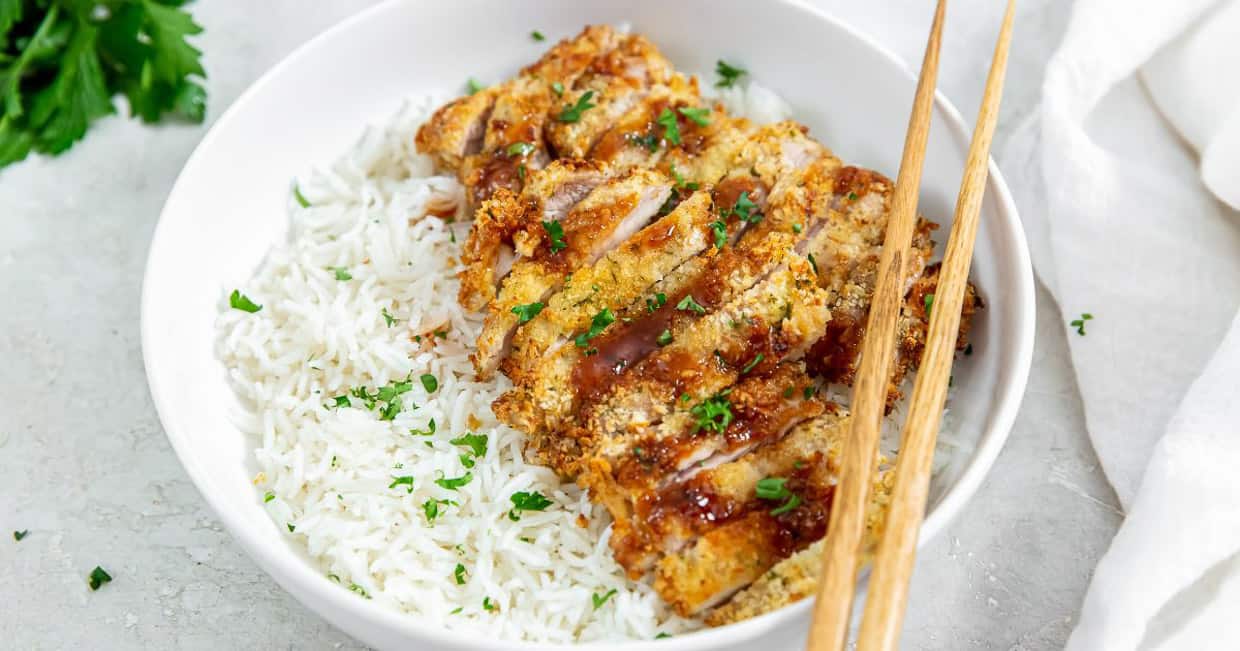 Air Fryer Katsu Chicken Thighs on a white plate with white rice, a brown sause, and parsley.