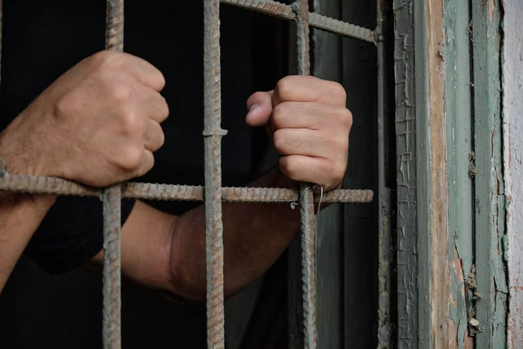 A caucasion man's hands holding prison bars from inside the cell.