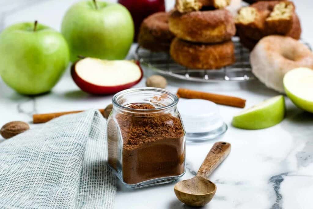 Apple pie spice in front of green apples and a baking rack with donuts.