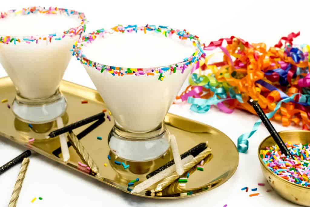 Two sprinkle-rimmed birthday cake martinis served on a gold tray next to curly ribbons, birthday candles and more colorful sprinkles.