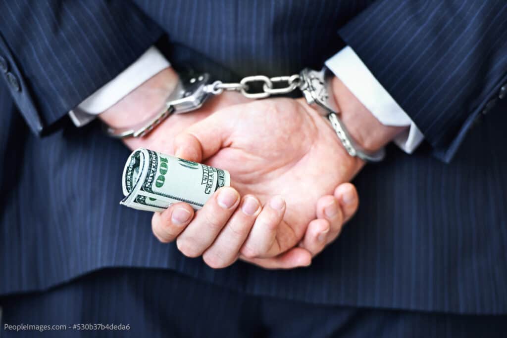 A businessmans hands in handcuffs and holding rolled up $100 bills, shot from behind.