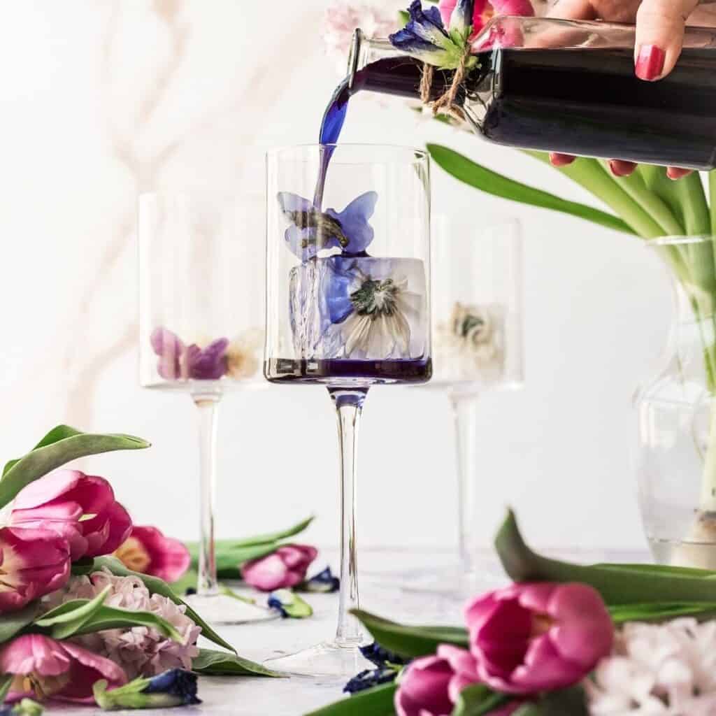 A purple liquid being poured into a tall clear glass filled with flowers.