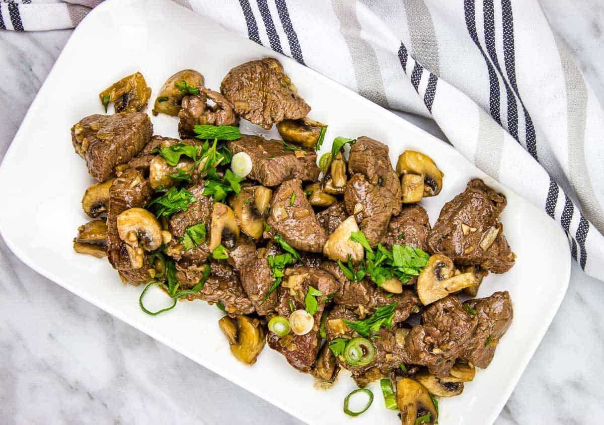 A white plate with garlic steak bites and mushrooms on it.