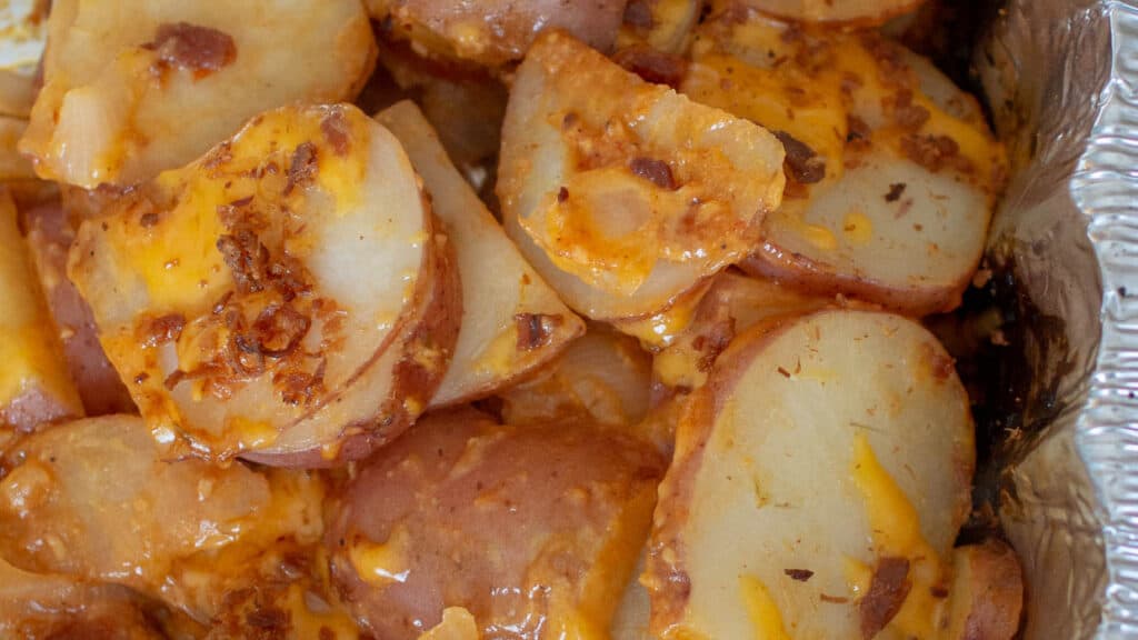 Grilled potatoes topped with cheese and bacon in a foil pan.