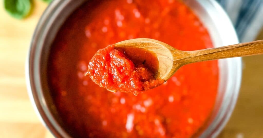 A closeup of hearty marinara sauce in a wooden spoon over a pot of finished marinara sauce.