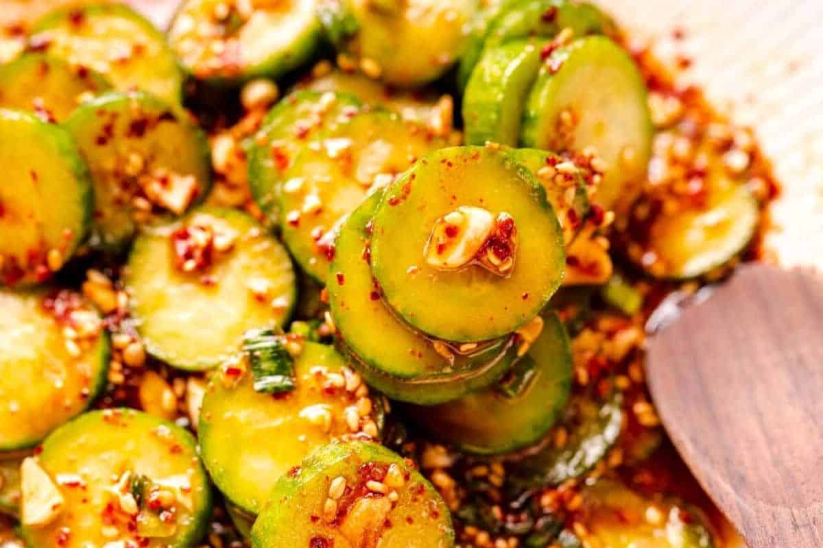 Cucumber slices coated in a red dressing in a white ceramic bowl with a wooden spoon.