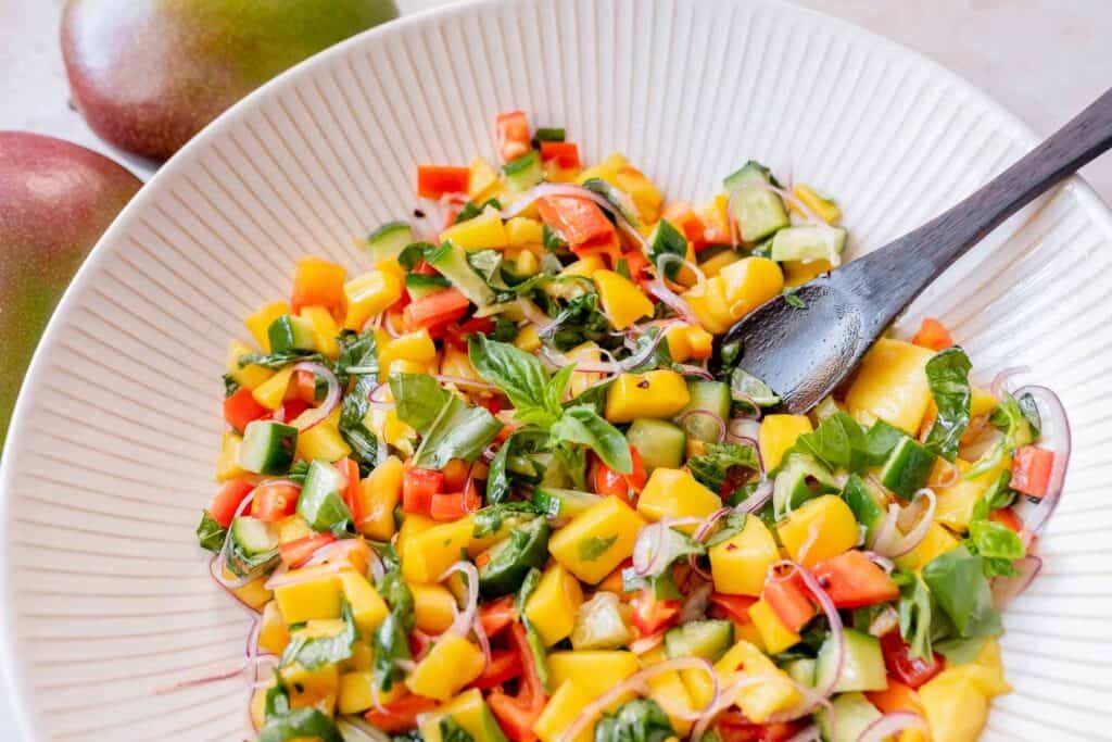 A colorful mango salad in a large ceramic bowl with a wooden spoon.