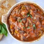 Top view of a big pot of mushroom stew with cheese and basil on a cutting board next to the pot.