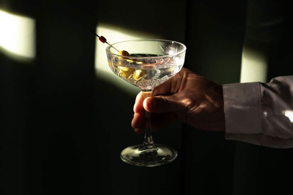 A hand holding a gin martini in a beam of sunlight surrounded by darkness. 