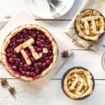 Three pies on a white wooden table, each with a crust featuring the Pi symbol&mdash;perfect for pi day activities. Two are small, one is large and topped with cherries. Plates, utensils, spices, and a bowl of cherries complete the festive scene.