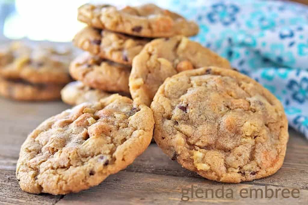 A stack of five potato chip cookies with three more cookies leaning against the front of the stack.  There is a turquoise and white print fabric napkin in the right background and two more cookies in the left background.  All cookies are on brown paper.