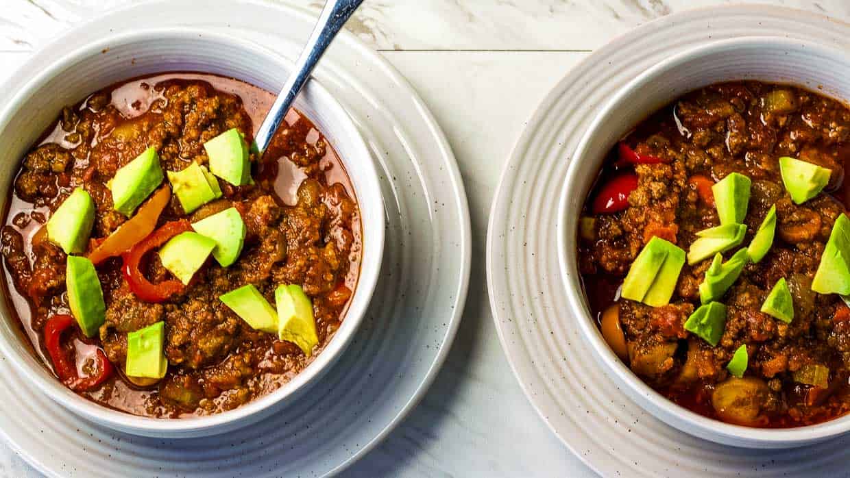 A slow cooker dinner recipe featuring two bowls of chili topped with avocado.