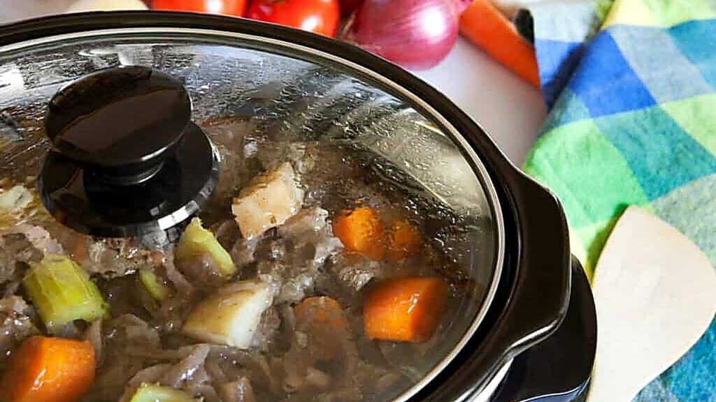 A slow cooker filled with a hearty stew of meat, carrots, potatoes, and celery sits on the counter near fresh vegetables and a checkered towel, showcasing the warmth and convenience that slow cookers bring to home-cooked meals.