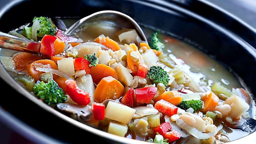 A ladle scoops a colorful vegetable soup with carrots, broccoli, red peppers, celery, and potatoes from a black pot&mdash;perfect for preparing in slow cookers for rich flavor and convenience.