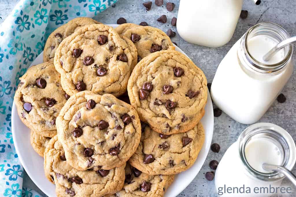 Small Batch Chocolate Chip Cookies served on a white plate. Plate and three clear glass bottles of milk are sitting on a concrete countertop next to a blue print fabric napkin.