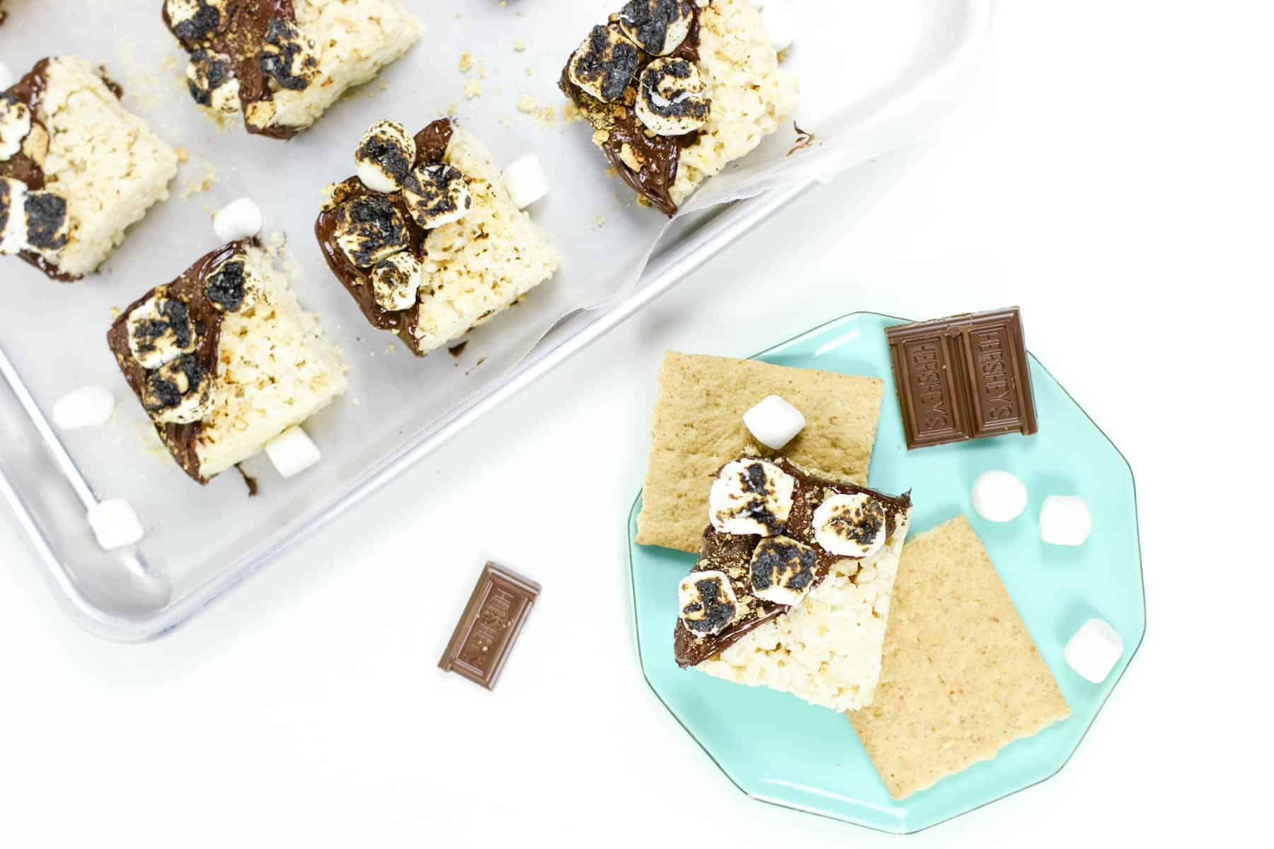 A tray of s'mores rice krispie treats next to a plate of the same. Each treat is dipped diagonally in chocolate and covered in toasted mini marshmallows and graham cracker crumbs.