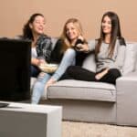 Three young women sitting on a couch, watching TV together&mdash;one holding a remote and another with a bowl of popcorn&mdash;enjoying one of the classic activities for teens.