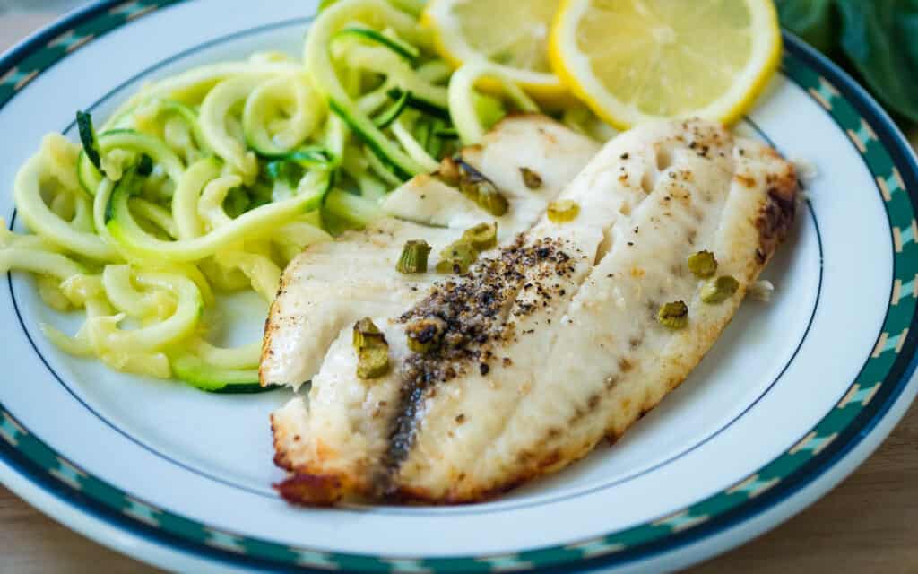 air fryer tilapia fillet on a plate with zucchini and lemon.