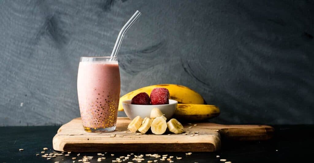 A strawberry banana oat smoothie with glass straw in front of strawberries and bananas on a wooden board, on a dark blue background with text overlay.