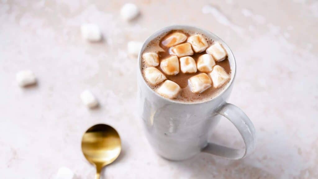 A white mug of hot cocoa with marshmallows rests on a white table next to a gold spoon.