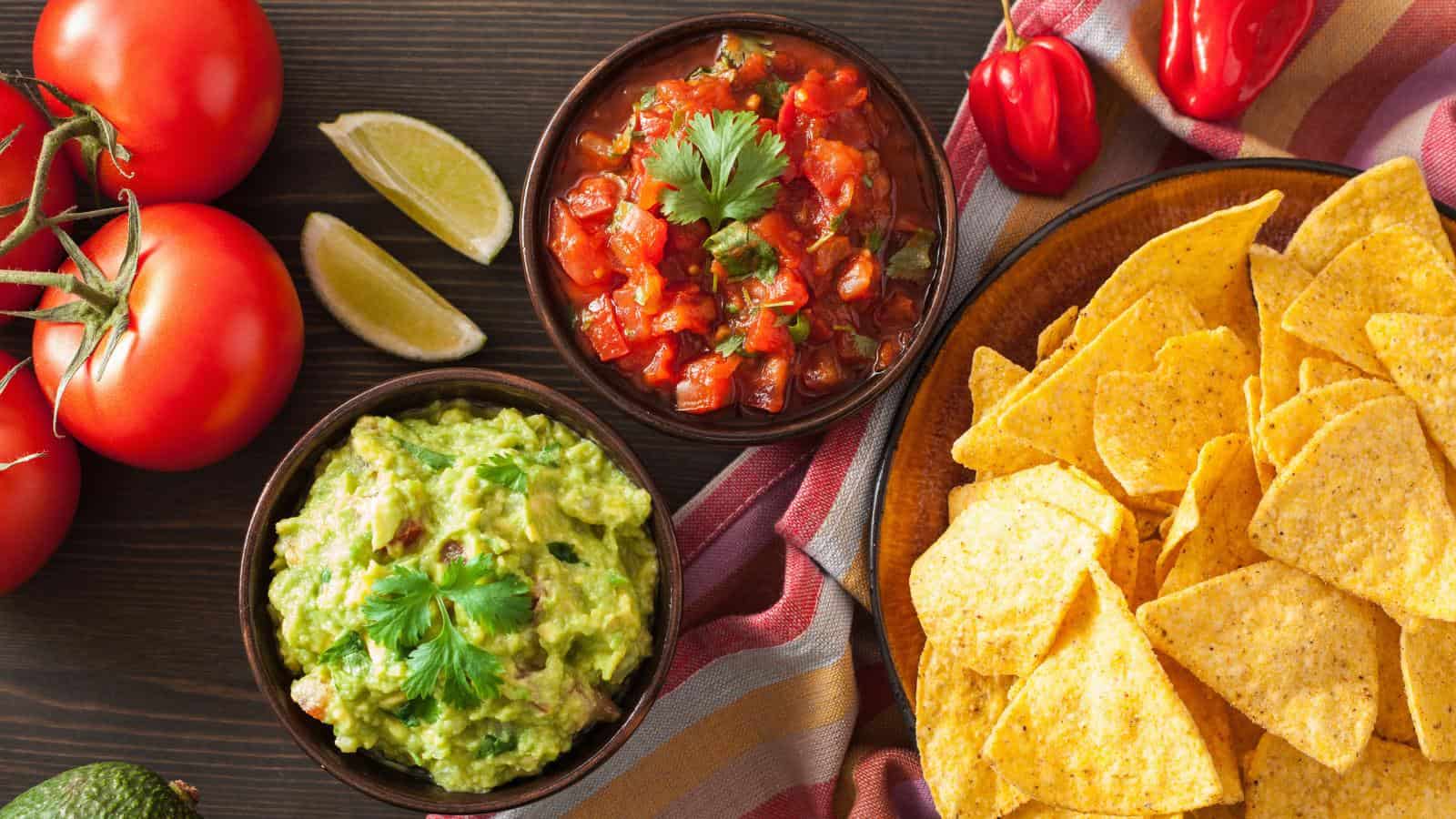 Two bowls of guacamole on tray with chips and various ingredients.
